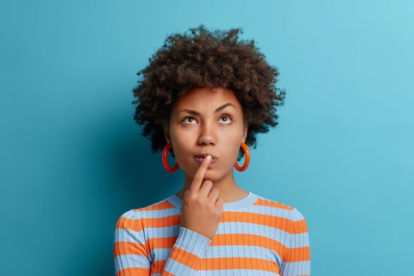 Vertical shot of thoughtful dark skinned woman has idea in mind, keeps finger on lips, looks pensively above, tries to decide how to act, poses against blue background, blank copy space above